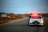 A policeman leans against a police car on an outback road as dusk approaches.