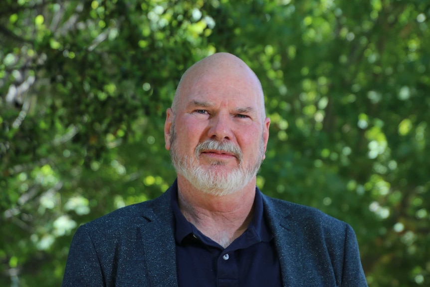 A bald bearded man wearing blue looks at the camera