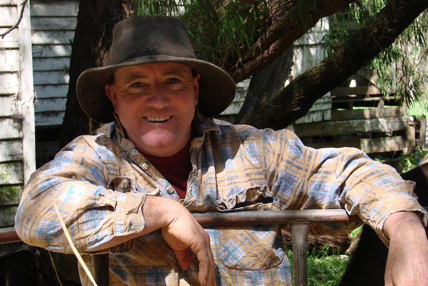 A smiling middle-aged farmer in a hat leans on a gate