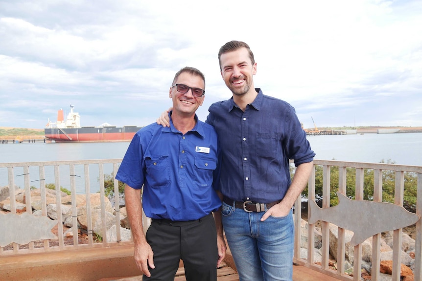 Two men stand on decking with a large ship at port behind