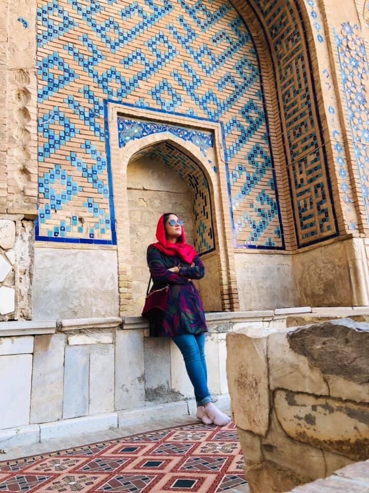 A young woman in jeans and a bright pink head scarf leaning against a mosque wall 