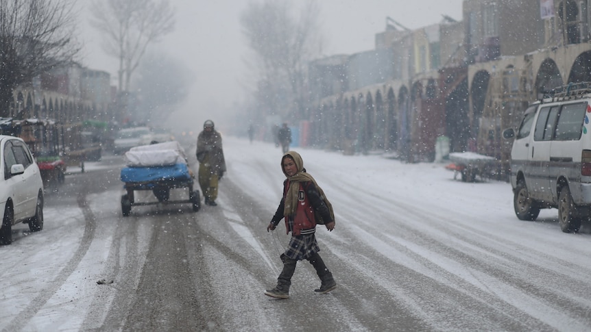 A girl crosses a snow covered road as a man pulls a cart in the background.