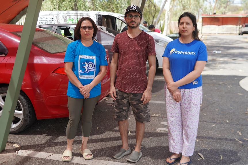 Two women and a man stand in a carpark in front of cars with smashed windows