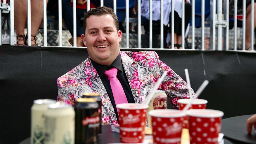 Man wearing punk floral suit smiles from behind a table with drinks at the Melbourne Cup.
