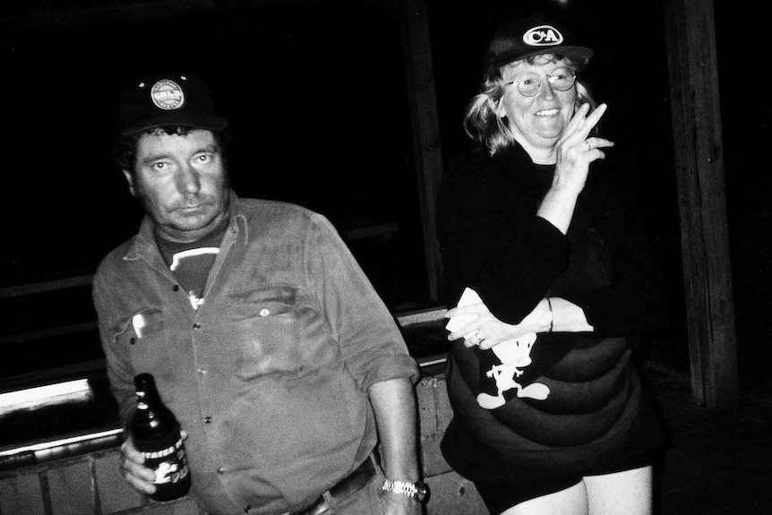 A black-and-white photo of a man holding a beer, standing beside a woman in glasses smoking a cigarette.