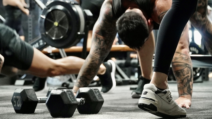 A man rests with his hands on the floor of a gym, with dumbbells near him and a woman walking past.