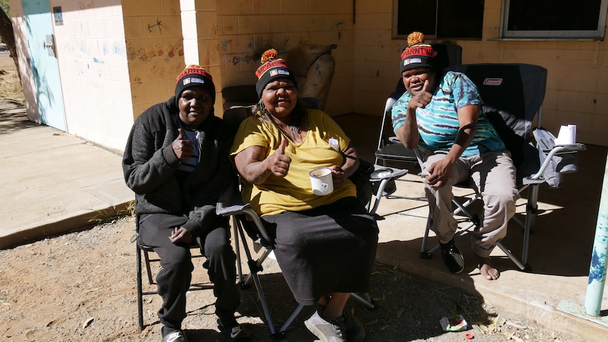 Three Indigenous women sitting in camp chairs and wearing matching beanies smile at the camera and give thumbs up