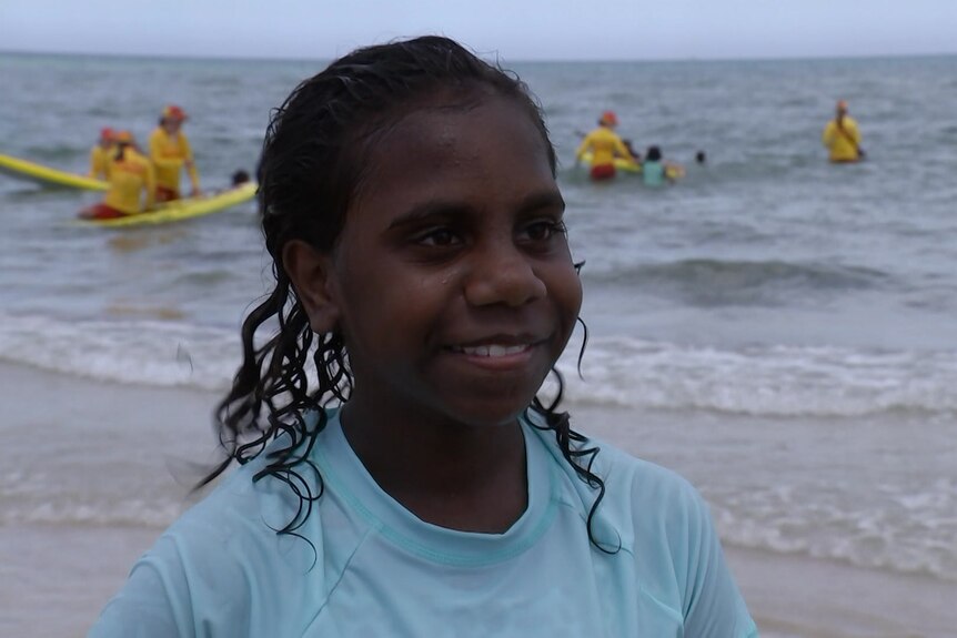 A girl wearing a blue rash vest stands on the shore line of a beach. In the background are surf life savers