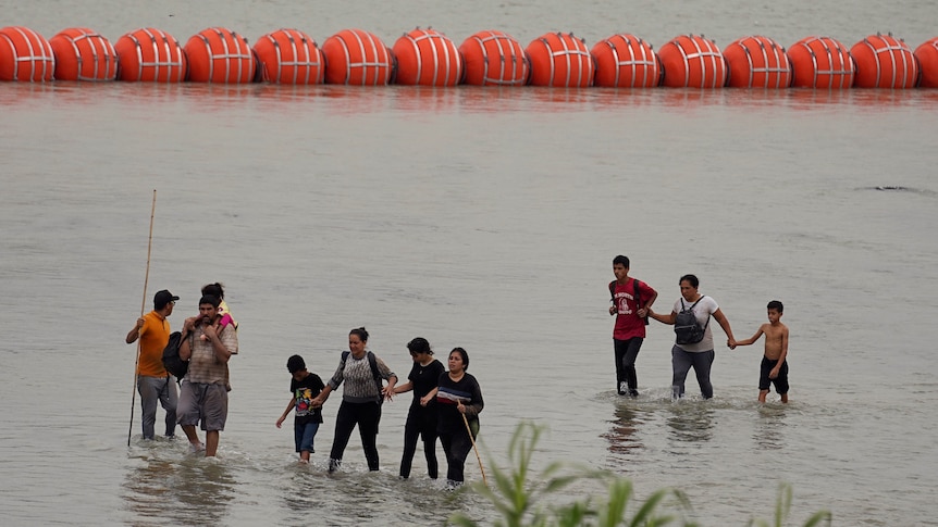 A group of people walk in shallow water in front of large orange floating buoys.
