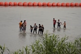A group of people walk in shallow water in front of large orange floating buoys.