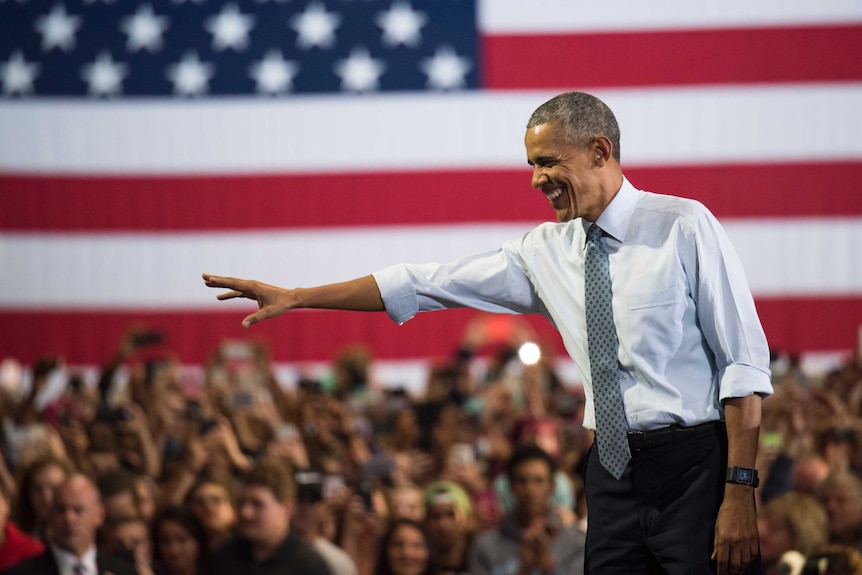Barack Obama waves to a crowd of people standing in front of a large American flag