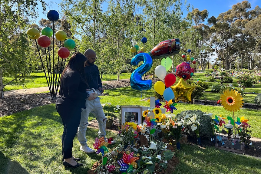 A man and a woman stand by a child's grave surrounded by colorful balloons and decorations.