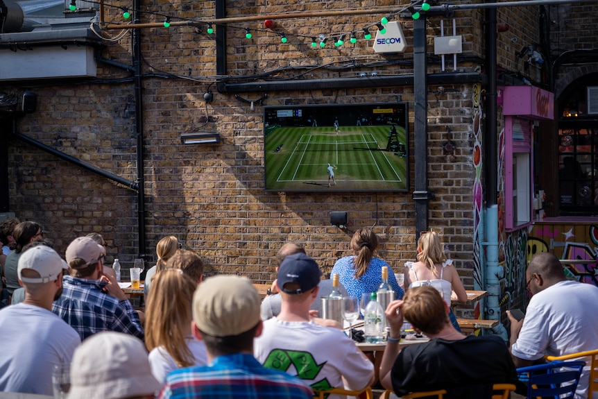 A group of people sit in a pub garden and watch the Wimbledon final on a television.