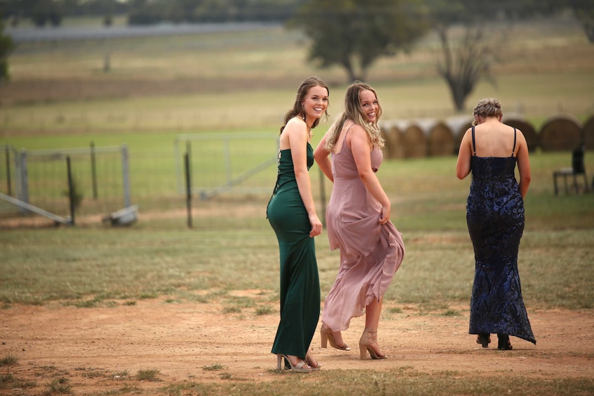 Three young women wearing colourful ball gowns walk along a dusty farm road, two look back smiling.