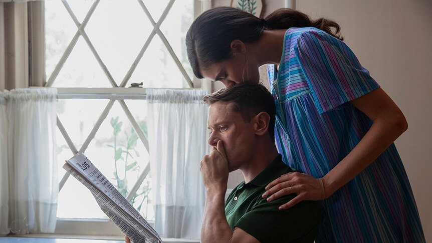 A man with shocked expression holds hand to mouth and reads newspaper next to kitchen window as a woman kisses top of his head.