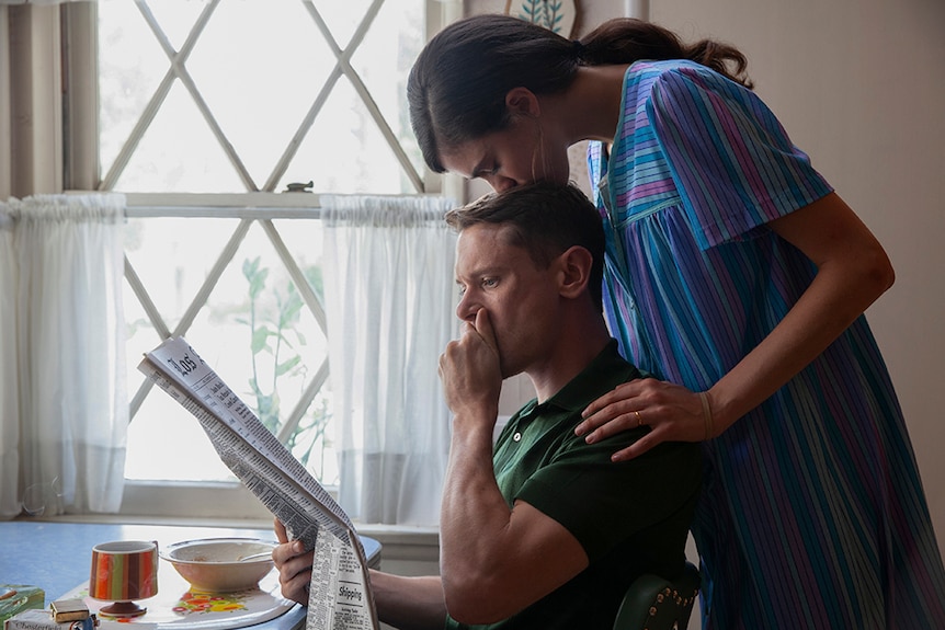 A man with shocked expression holds hand to mouth and reads newspaper next to kitchen window as a woman kisses top of his head.