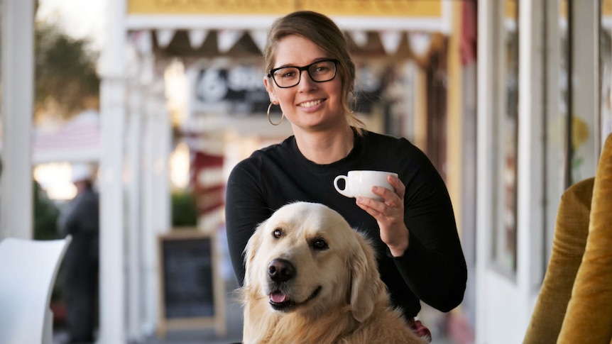 A woman with dark blonde hair and glasses sitting on an armchair outside a cafe, with a golden retriever dog.