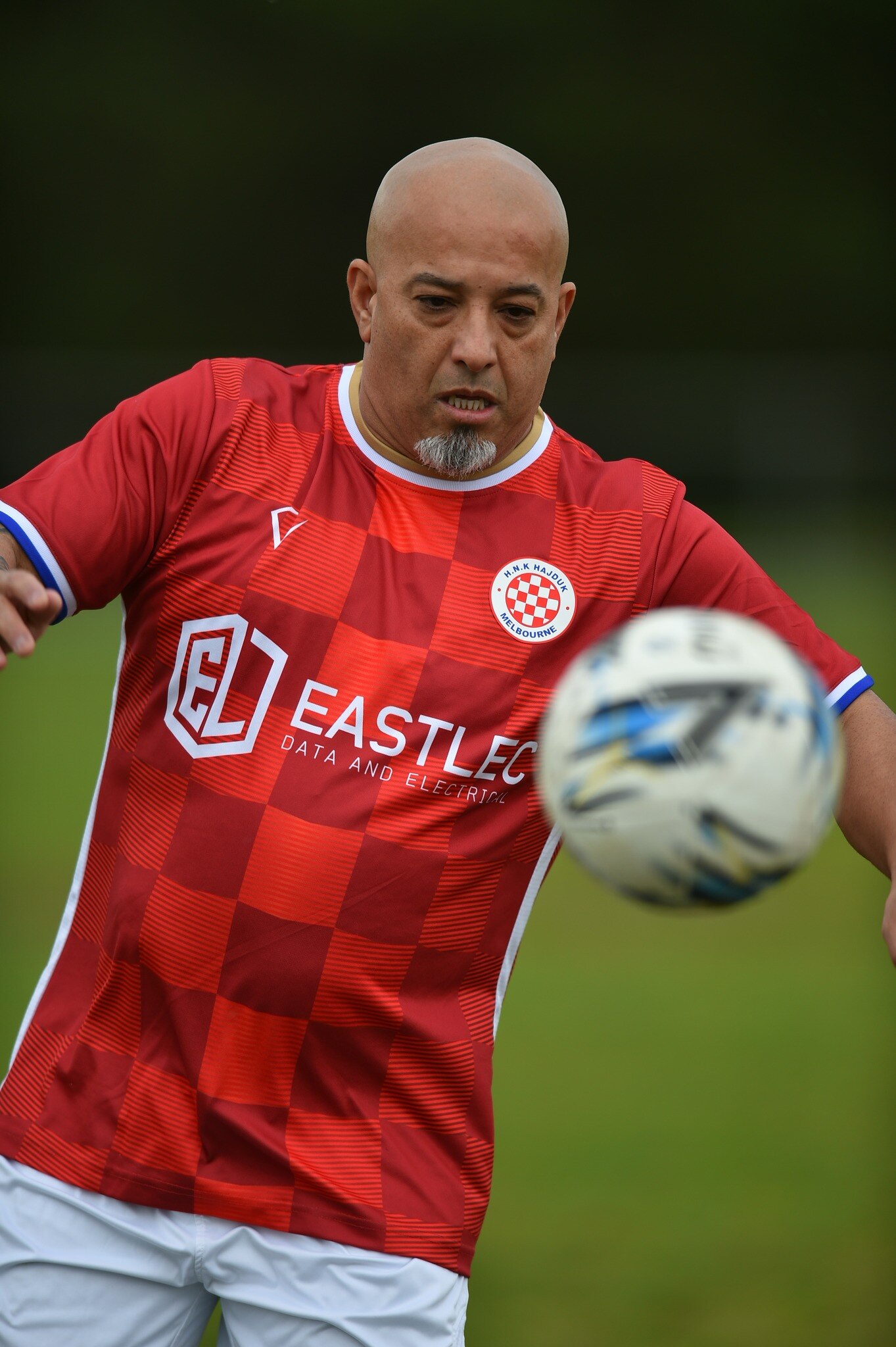 A soccer ball hangs in the air in front of a bald man in a red soccer shirt and white shorts.