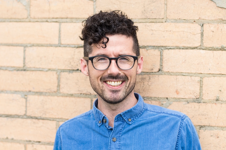 Caleb, wearing a blue button-up shirt and glasses, smiles as he stands in front of a light-brown brick wall.