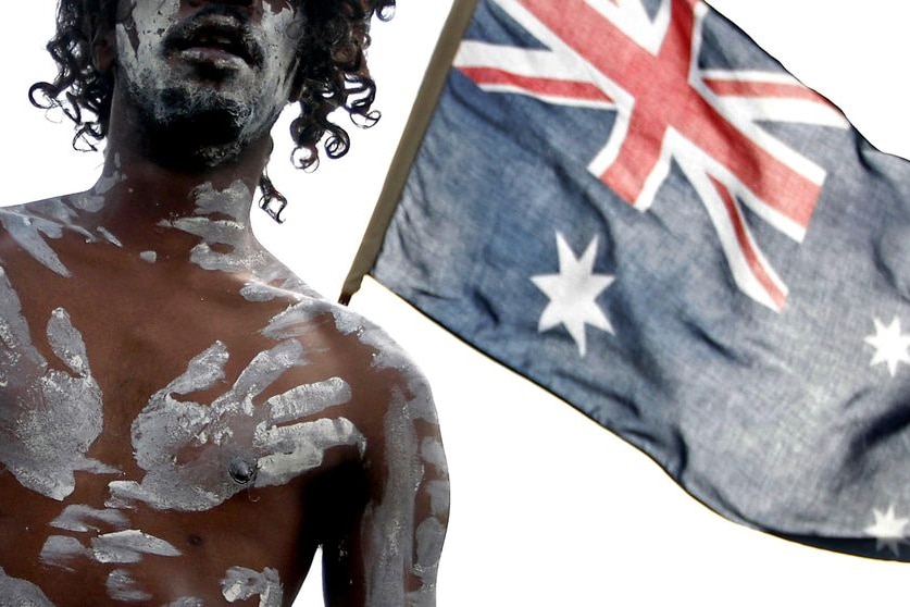 An Aboriginal performer stands in front of an Australian flag