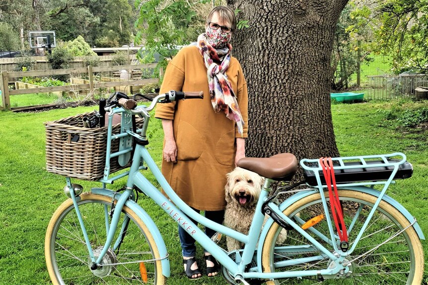 A woman with short brown hair wears a long top, scarf and facemask, she stands in a garden with the bike and her dog.