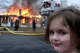 a young child turns her head to smile at the camera as a house burns down behind her.