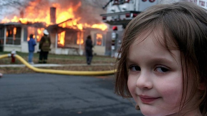 a young child turns her head to smile at the camera as a house burns down behind her.