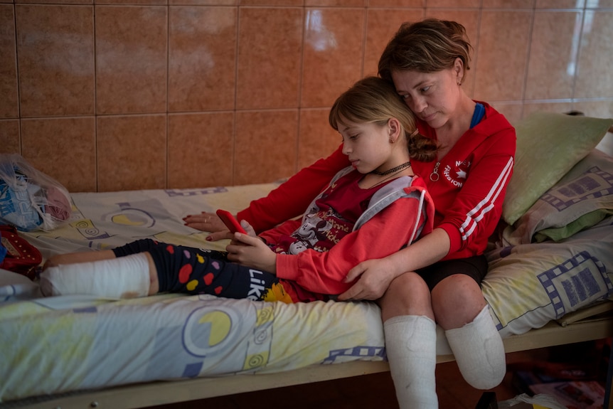 A girl with an amputated leg lies on a bed looking at a mobile phone, embraced from behind by a woman with two amputated legs