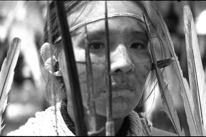 A black and white still from a video artwork featuring an androgynous person wearing tribal-like face paint and feathers.