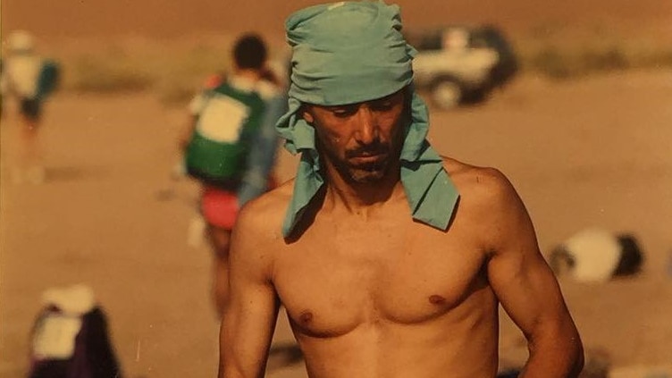 A shirtless man with a blue cloth tied around his head, running through the desert 