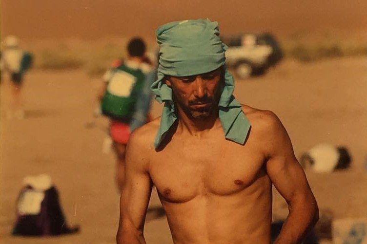 A shirtless man with a blue cloth tied around his head, running through the desert 