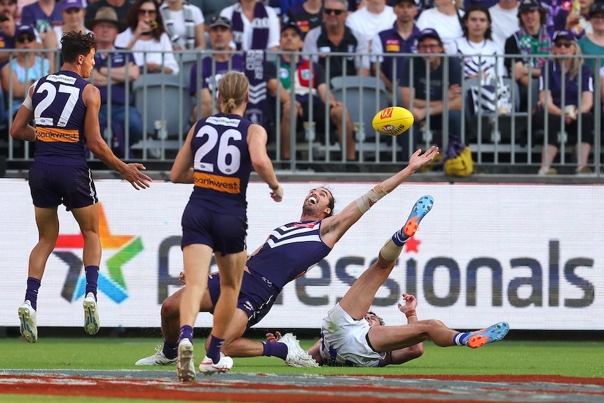 A Fremantle Dockers defender bends backwards and reaches to try and grab the ball, while Kangaroos forward lies on the ground.