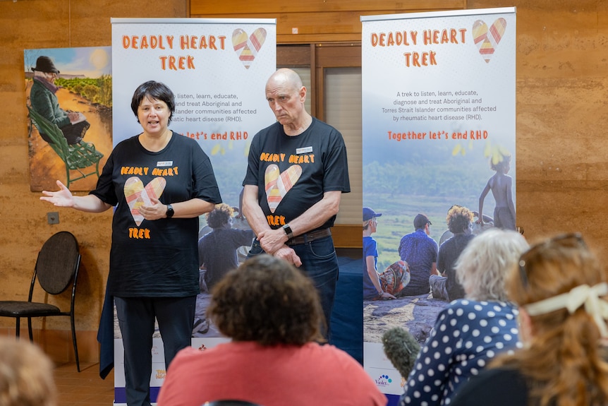 Two doctors, one woman and one older man, are standing in front of two posters and delivering a speech to a group. 