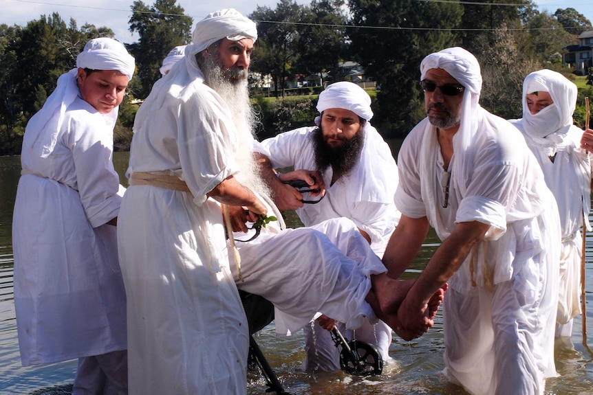 Men in white robes carry a elderly man in a wheelchair out of a river, after he has been baptised.