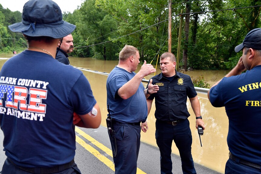 Grim-looking firefighters in blue shirts and trousers converse on a rural street partly flooded with brown water.