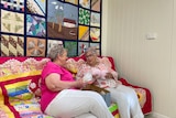 Two women sit on a brightly coloured holding knitted breasts, with a patchwork quilt on the wall behind them. 