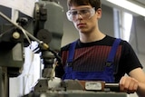 A teenage male wearing safety goggles stands by a welding machine