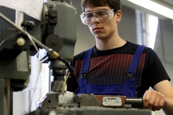 A teenage male wearing safety goggles stands by a welding machine