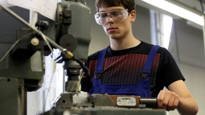 A teenage male wearing safety goggles stands by a welding machine