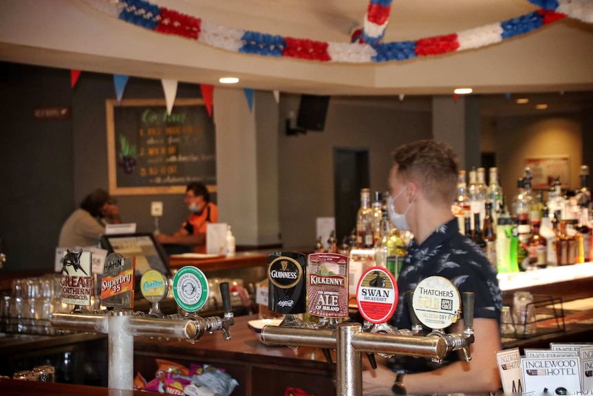 A man stands behind a bar at the Inglewood hotel with beer taps in the foreground and patrons in the background.