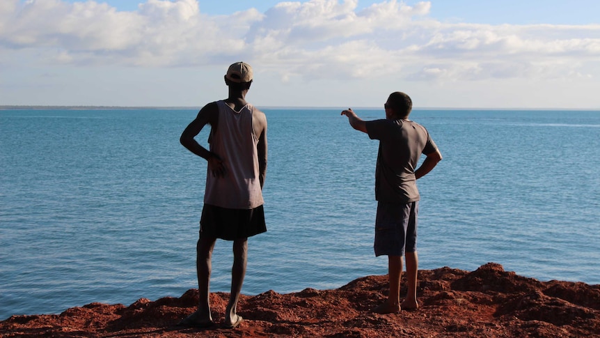 Wideshot of two men, one pointing to the horizon, standing on the edge of water.