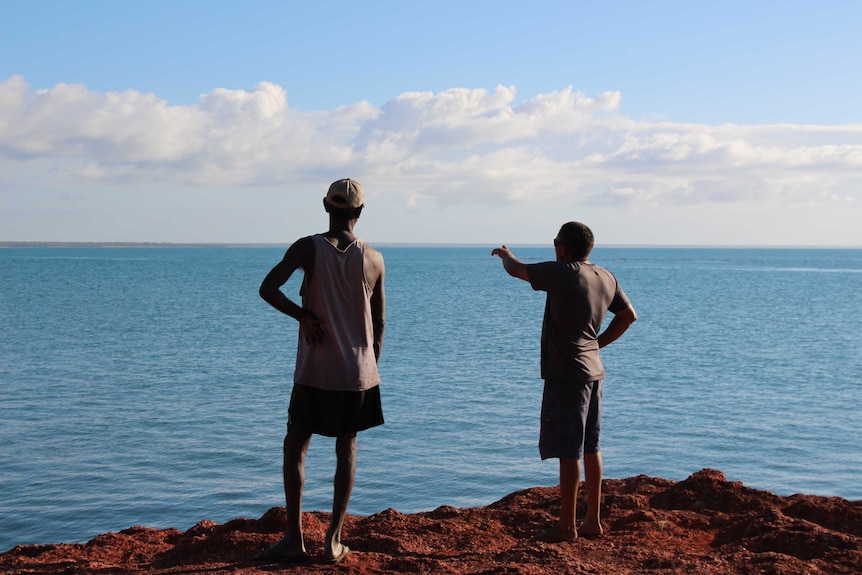 Wideshot of two men, one pointing to the horizon, standing on the edge of water.