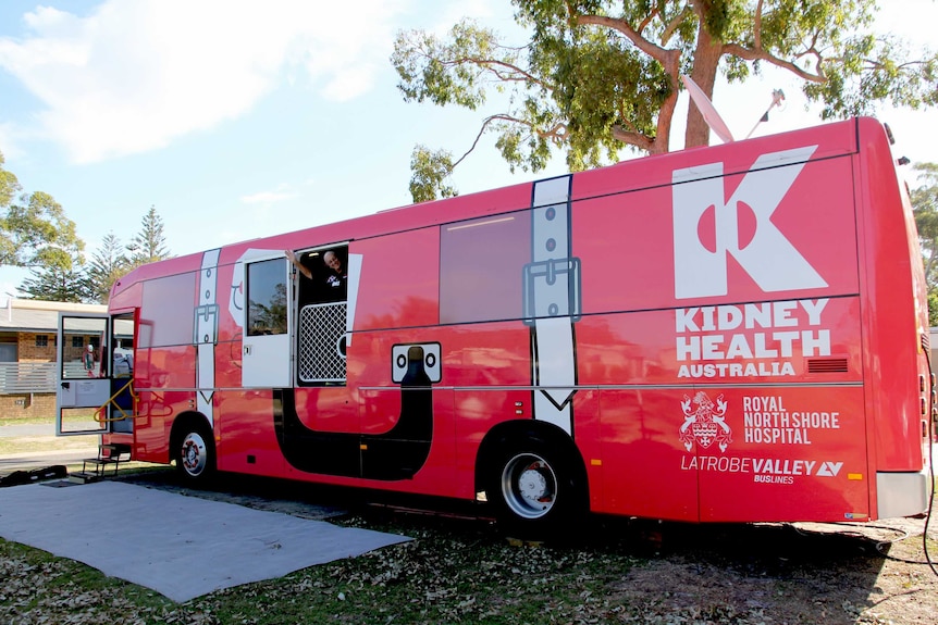 A big red bus is parked at a caravan park with a woman waving out a window