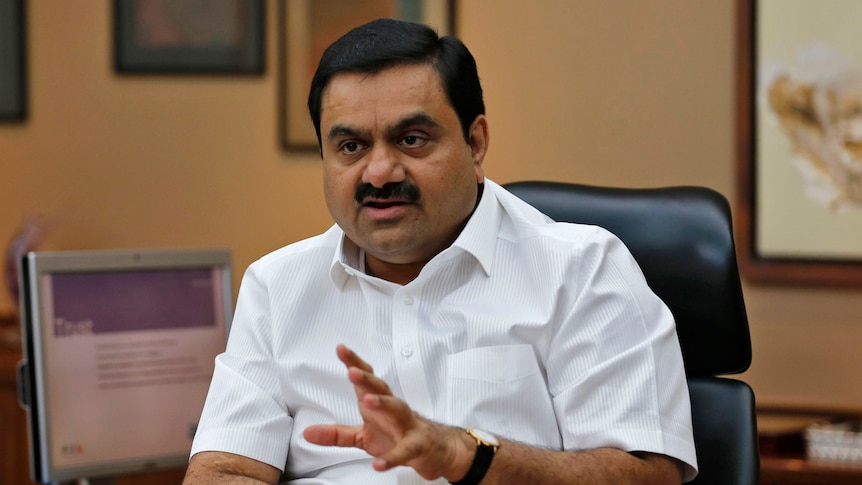 Mr Adani sits at a desk gesuring with his hands. 