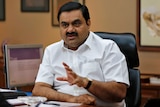 Mr Adani sits at a desk gesuring with his hands. 