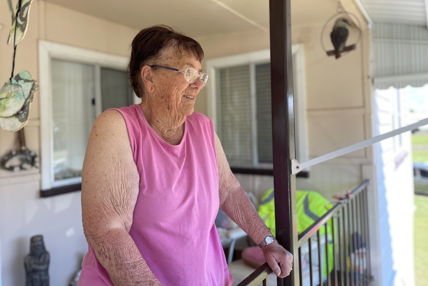 An elderly lady standing on a balcony smiling into the distance. 