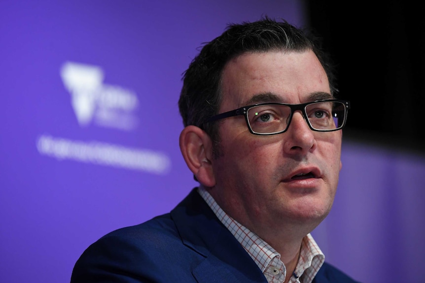 Daniel Andrews wears black-rimmed glasses and a blue suit in front of a purple backdrop with a blurred white logo.