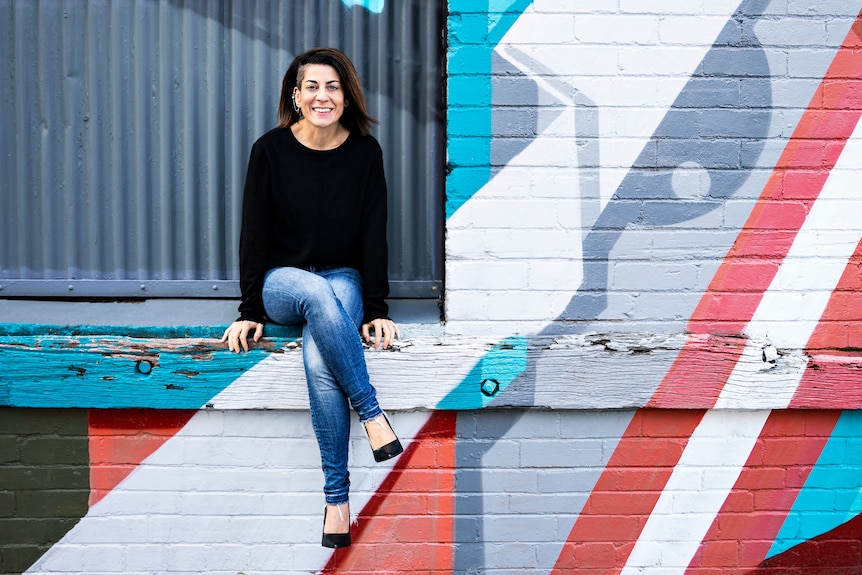 A woman with black hair and a black top sitting in front of a colorful mural. 