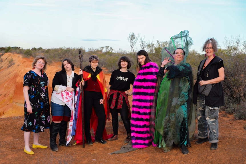 Six people stand on red dirt, some are wearing bright costumes, the sky is blue.