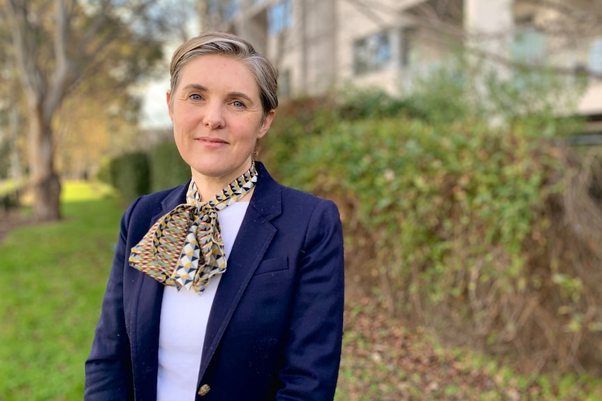 A woman wearing a white shirt, dark blue blazer and scarf standing in front of a hedge with buildings behind it. 